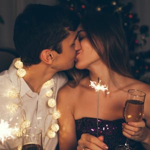 Christmas and New year party concept. Couple kissing, burning sparklers by illuminated Christmas tree with champagne.