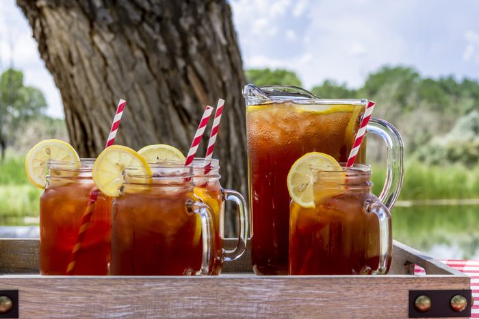 Pitcher and mason jar mugs filled with iced tea and lemons sitting on picnic table with red checked tablecloth