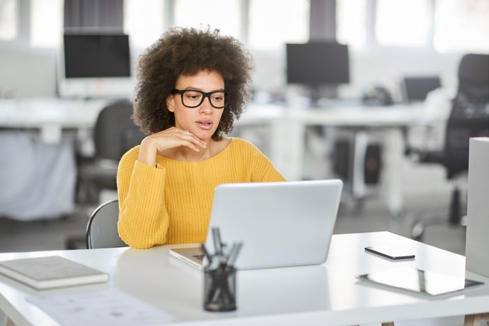Serious mixed race businesswoman dressed casual sitting in modern office and using laptop.