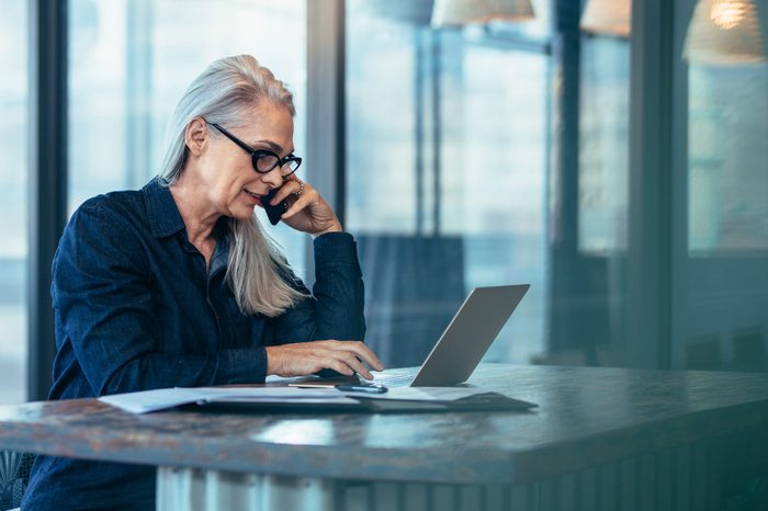 Senior business woman talking on cell phone while working on laptop in office. Mature female using laptop and talking on mobile phone.