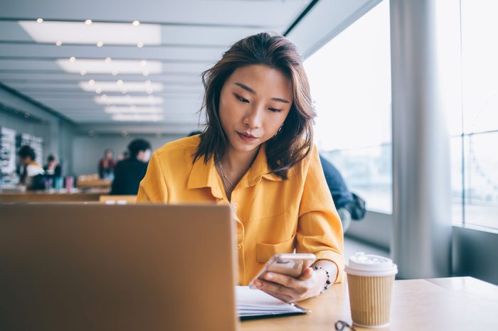 Beautiful focused Asian woman browsing smartphone in front of laptop in workspace in Hong Kong city with people in background