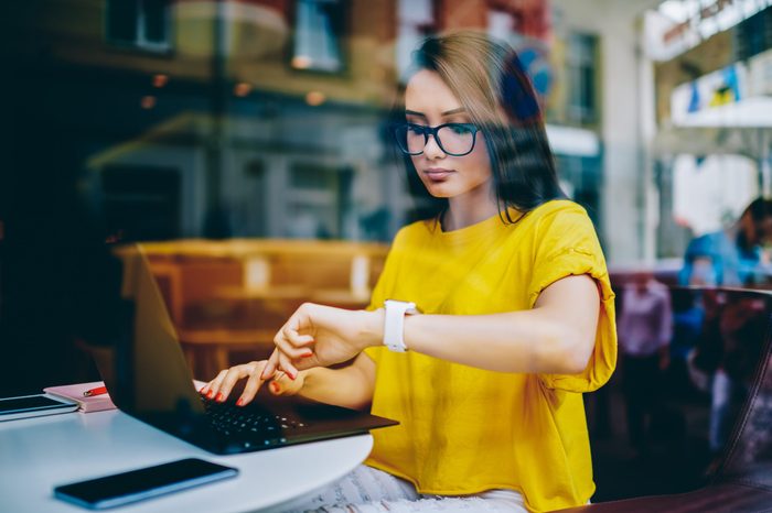 Young businesswoman in eyeglasses looking at display of wristwatch and managing time while checking email on laptop.Freelancer with smartwatch on hand working on article at netbook before deadline