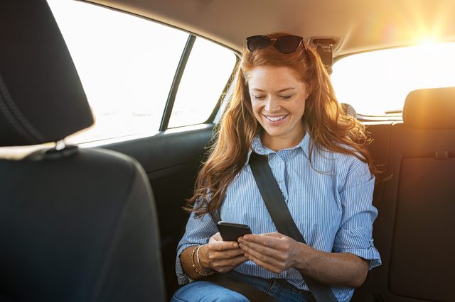 Smiling casual woman sitting in back seat using mobile phone. Cheerful young woman reading messages in smartphone while sitting in a taxi. Attractive girl with red hair wearing blue shirt in car using cellphone.