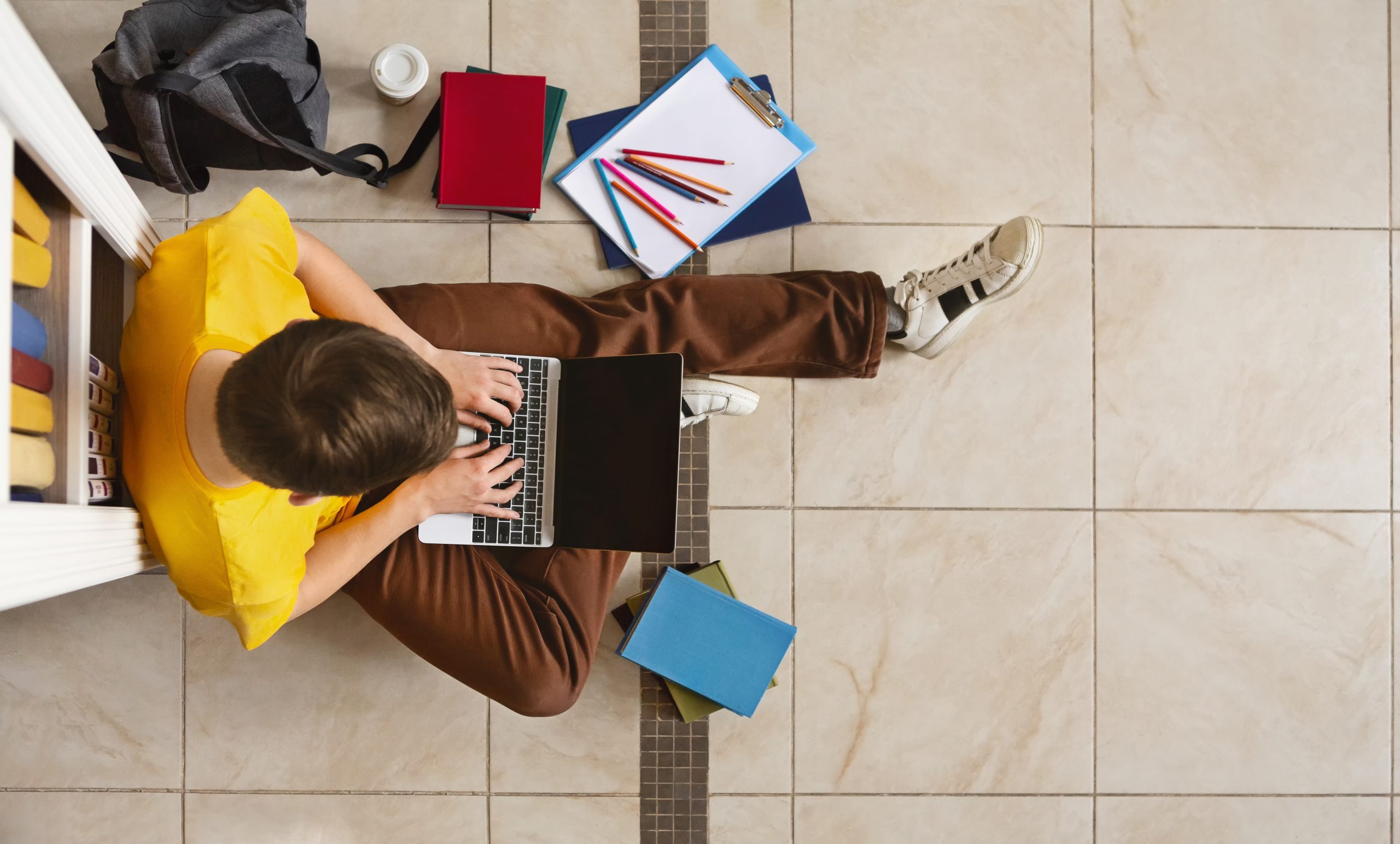 Young guy sitting next to bookshelf at library, using laptop