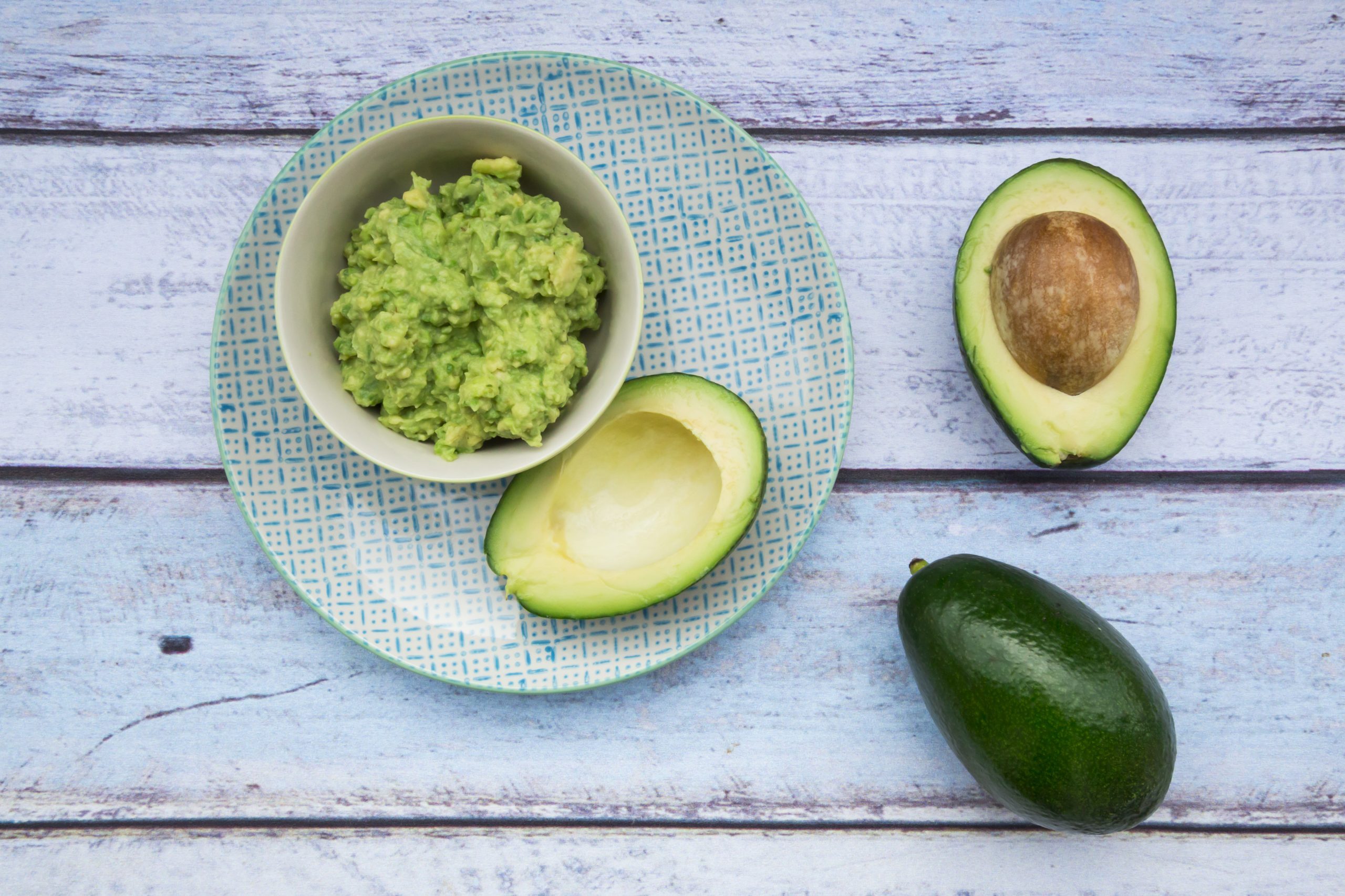 Bowl of Guacamole and whole and sliced avocados on light blue wood