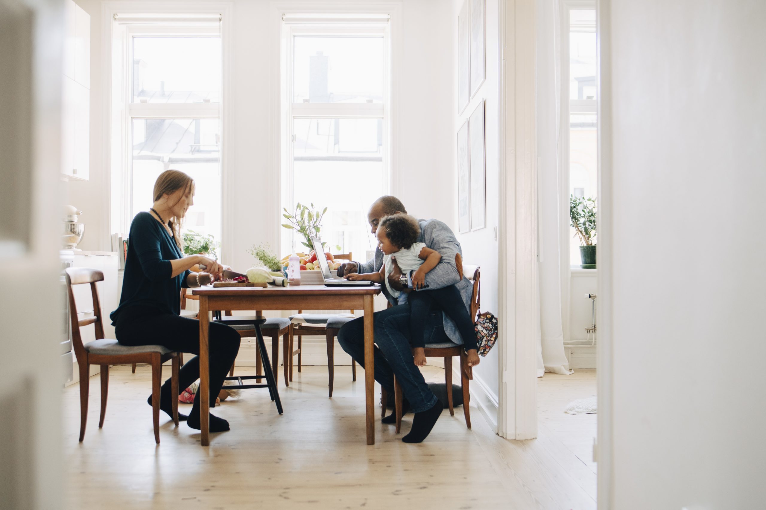 Father showing laptop to daughter while mother cutting vegetables at table in house