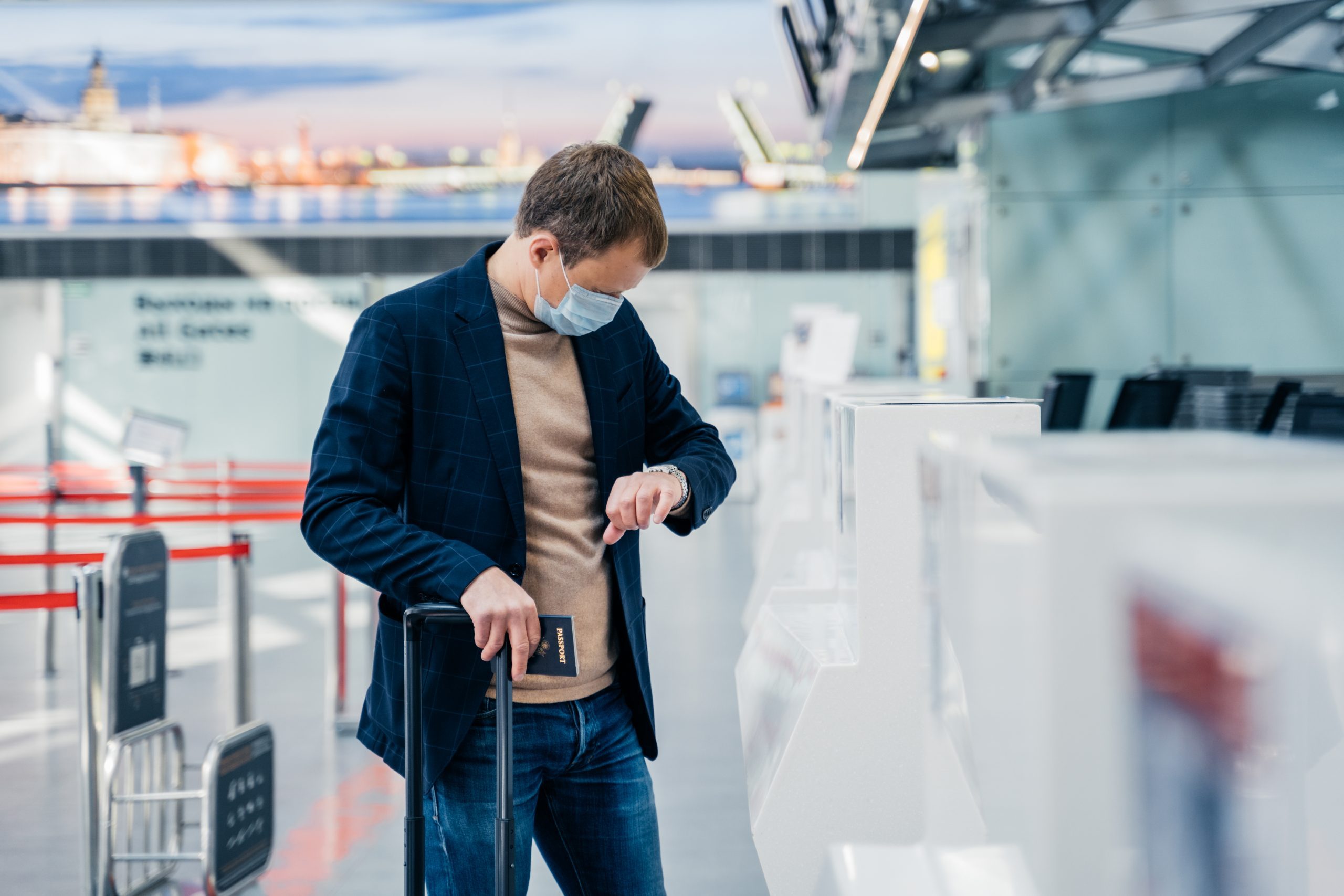 Horizontal shot of man passenger wears disposable medical mask to prevent virus, checks time on his watch, has hour for departure, poses in internation airport with suitcase and passport. Coronavirus