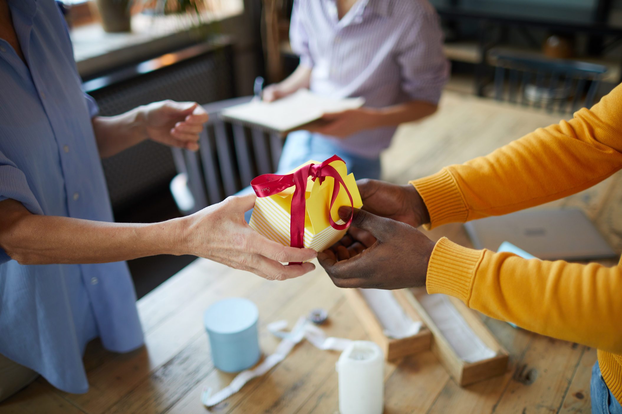 Close-up of unrecognizable craftspeople adjusting paper gift box in workshop: woman giving yellow box with red ribbon to black man