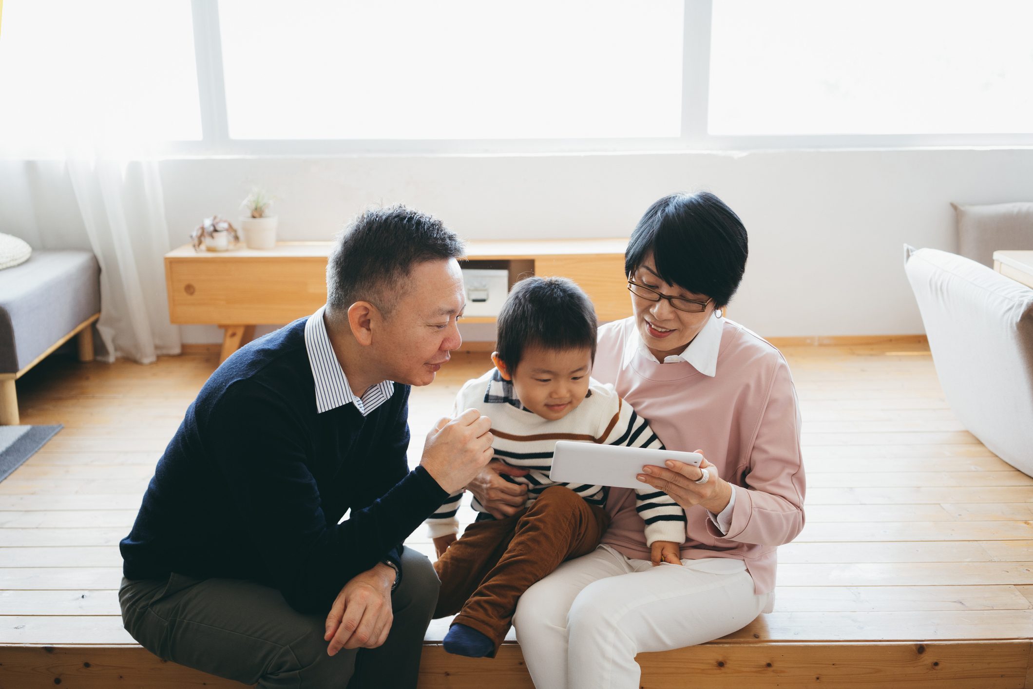 Joyful Asian grandparents sitting on the floor in the living room using digital tablet with little grandson together at home