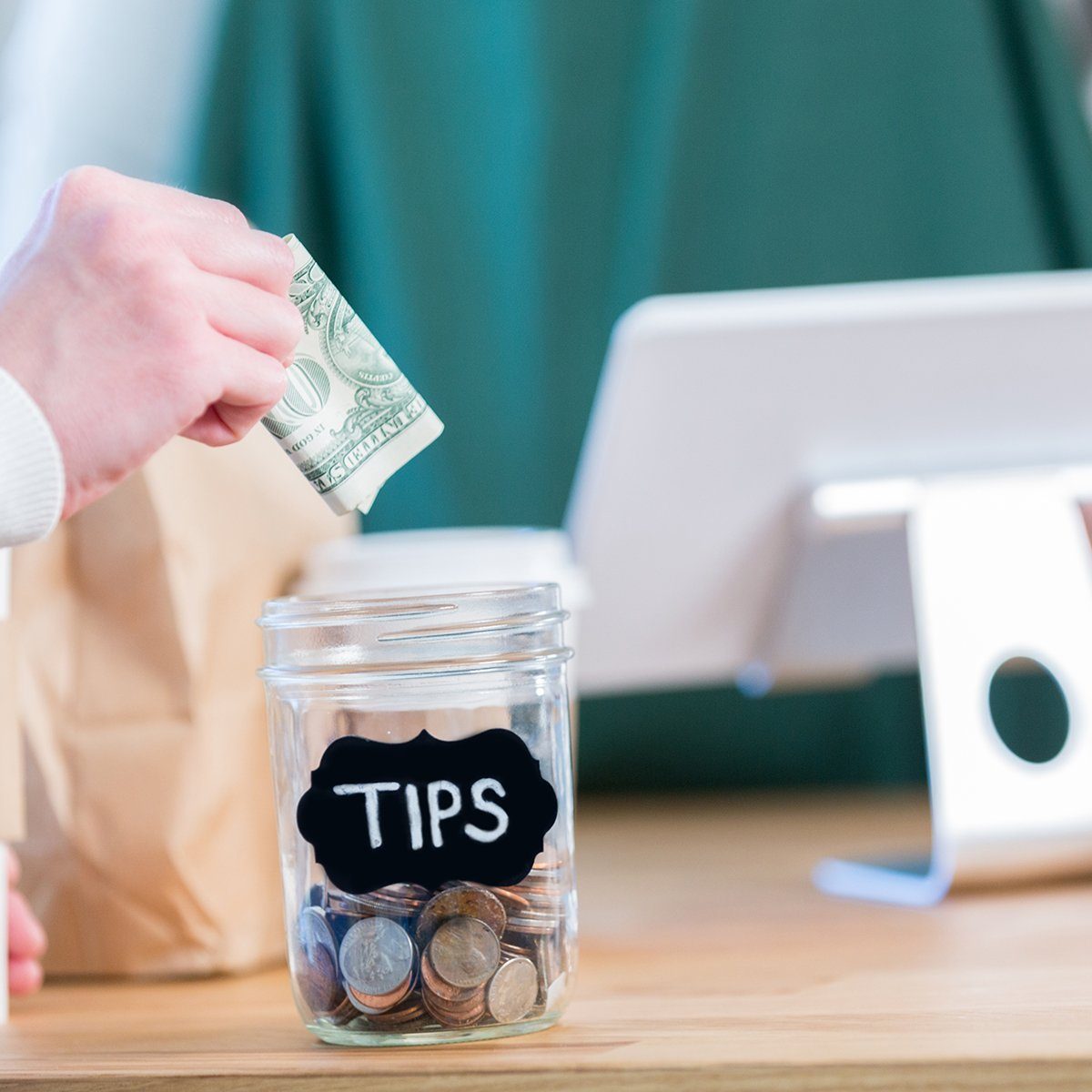 An unrecognizable coffee shop customer stands across the checkout counter from an unrecognizable barista and reaches out to put a paper bill in the tips jar.