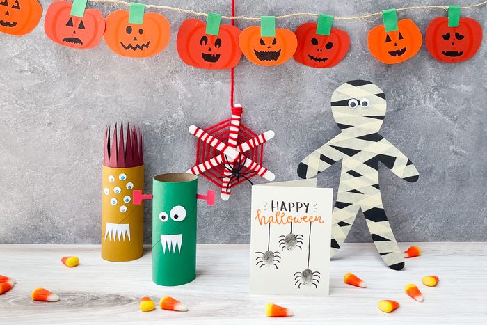 Kids Halloween Crafts arranged on a simple background; jack-o-lantern garland, toilet paper roll monsters, yarn and popsicle stick spider web, thumbprint spider card, masking tape mummy
