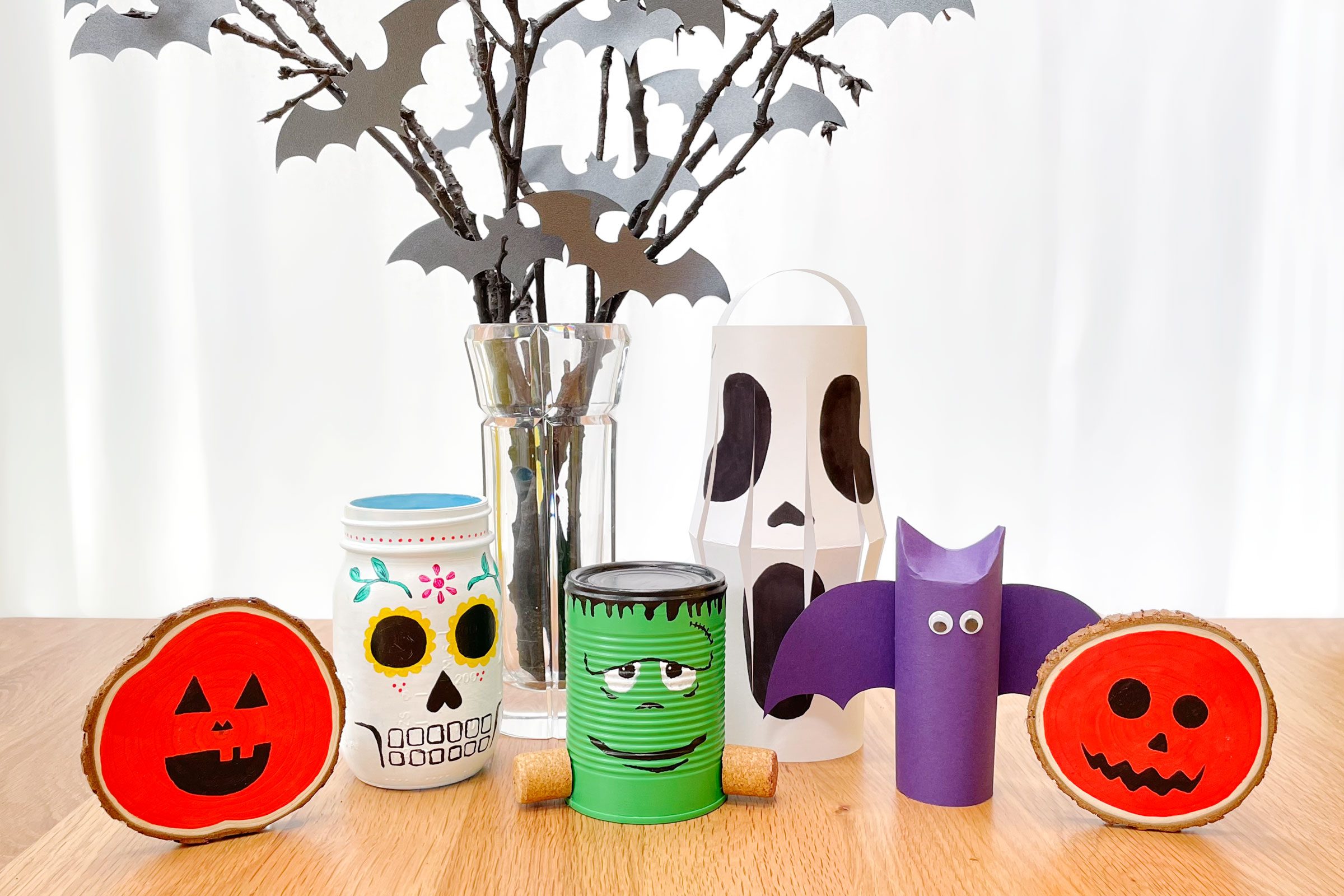 cute, cheap halloween decorations on a wooden table with white background; pumpkin wood slices, mason jar sugar skull, spooky centerpiece, tin can frankenstein, paper scream lantern, paper bat