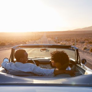 Rear View Of Couple On Road Trip Driving Classic Convertible Car Towards Sunset