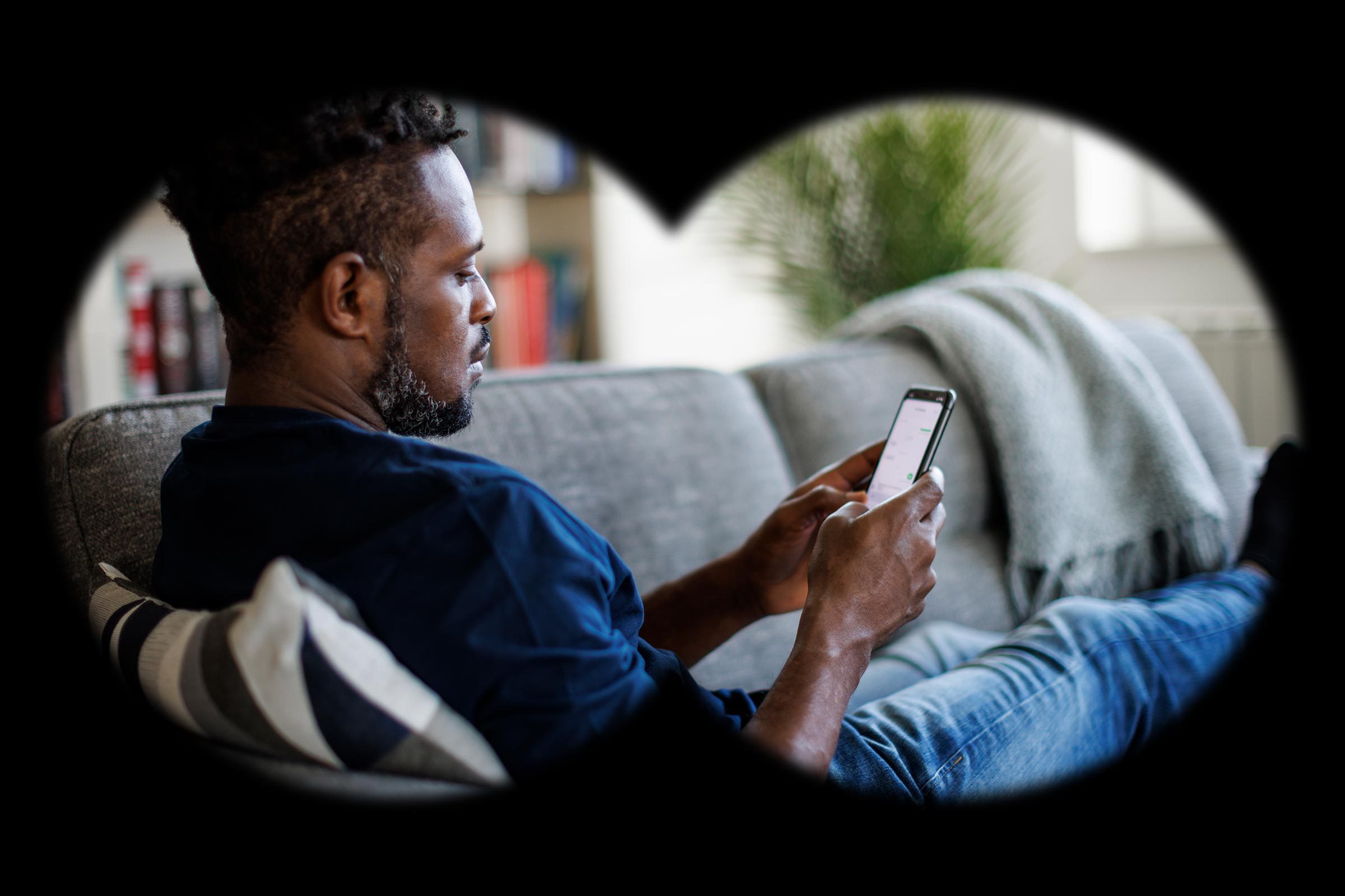 view from inside binoculars, from a spying point of view; man sitting on the couch at home using an iphone