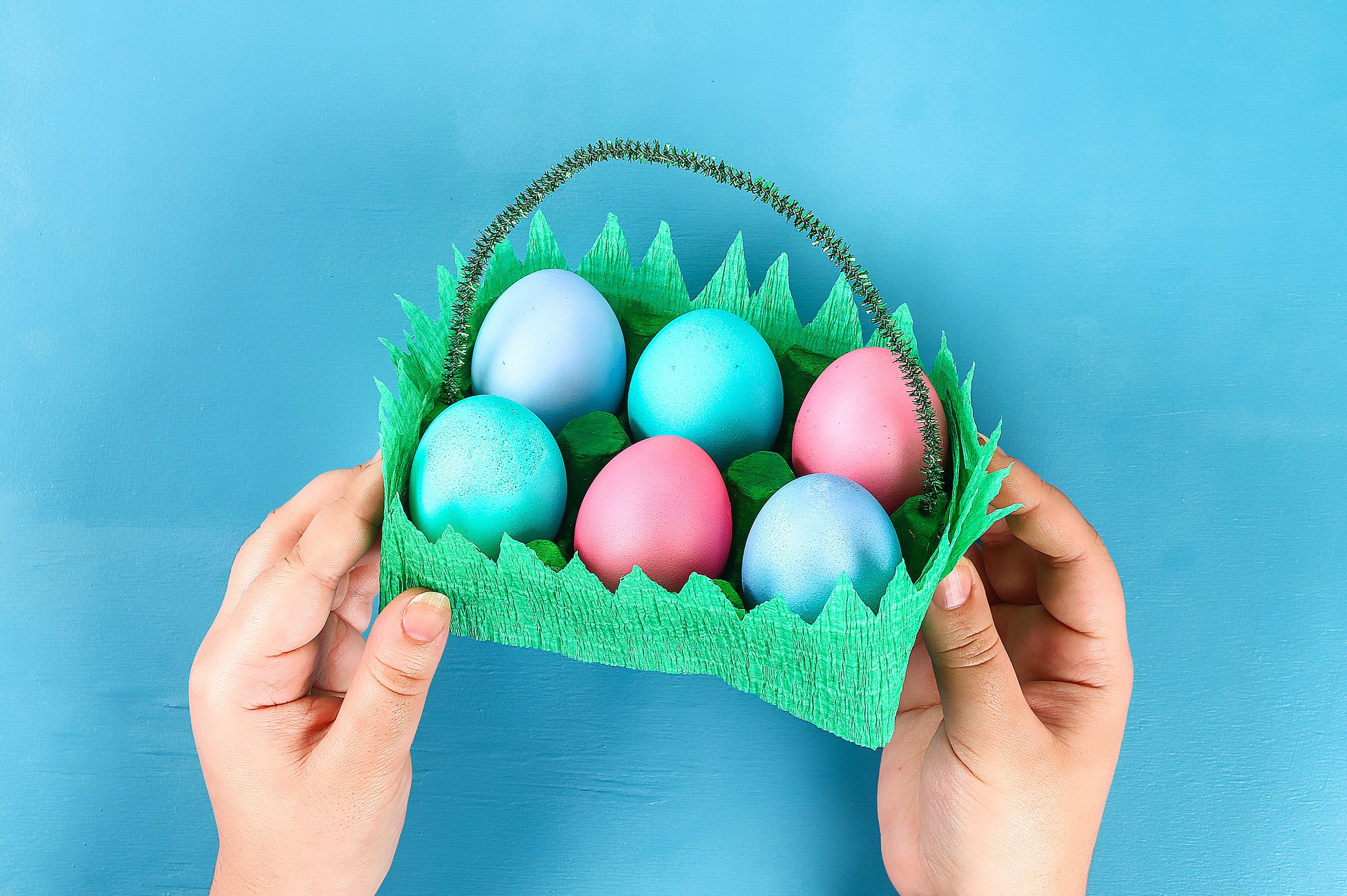 DIY basket Easter egg from cardboard tray, crepe paper, chenille stem on blue background.