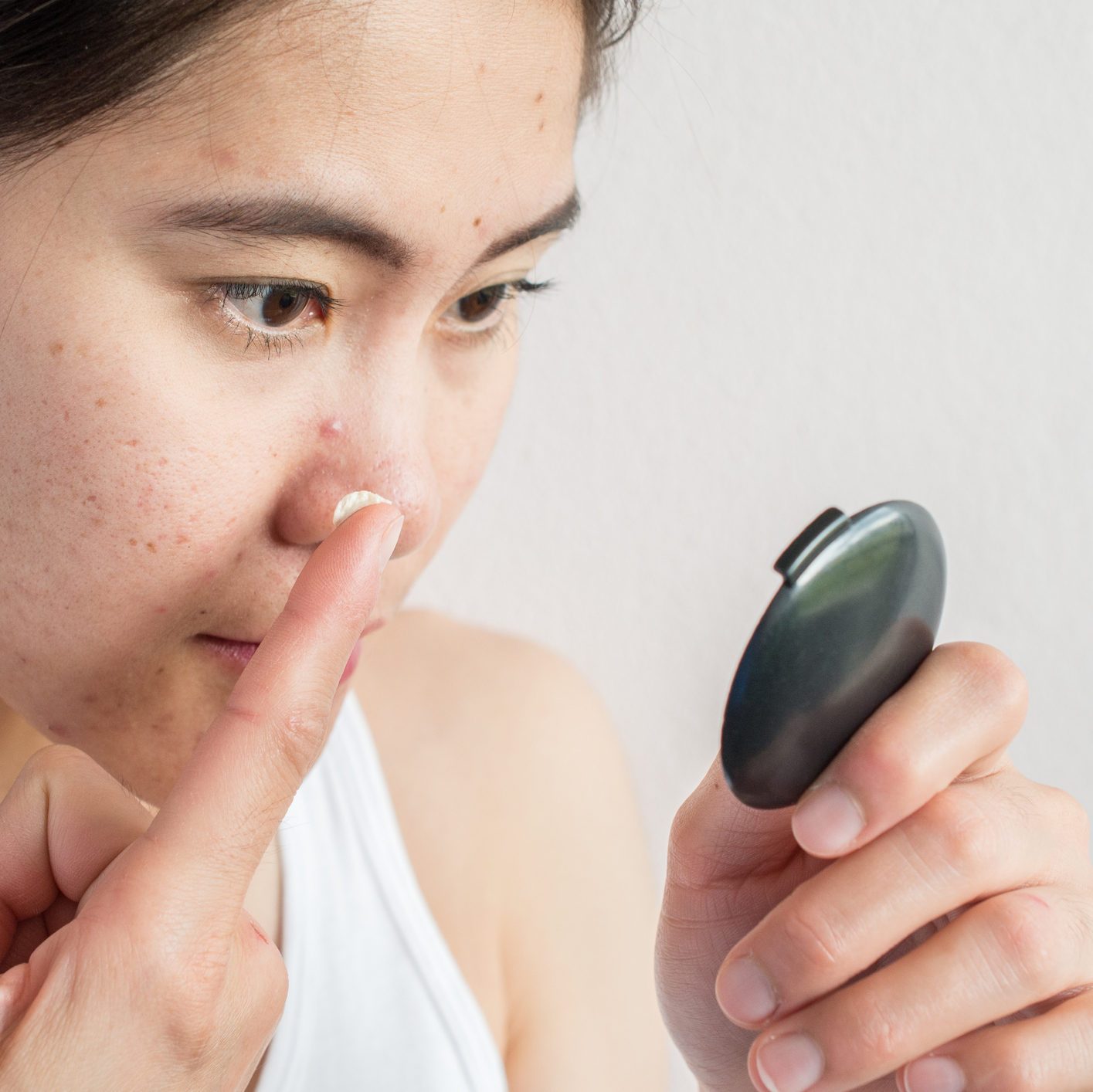 Portrait of young Asian woman applying acne cream/moisturizer on her face by looking a pocket mirror.