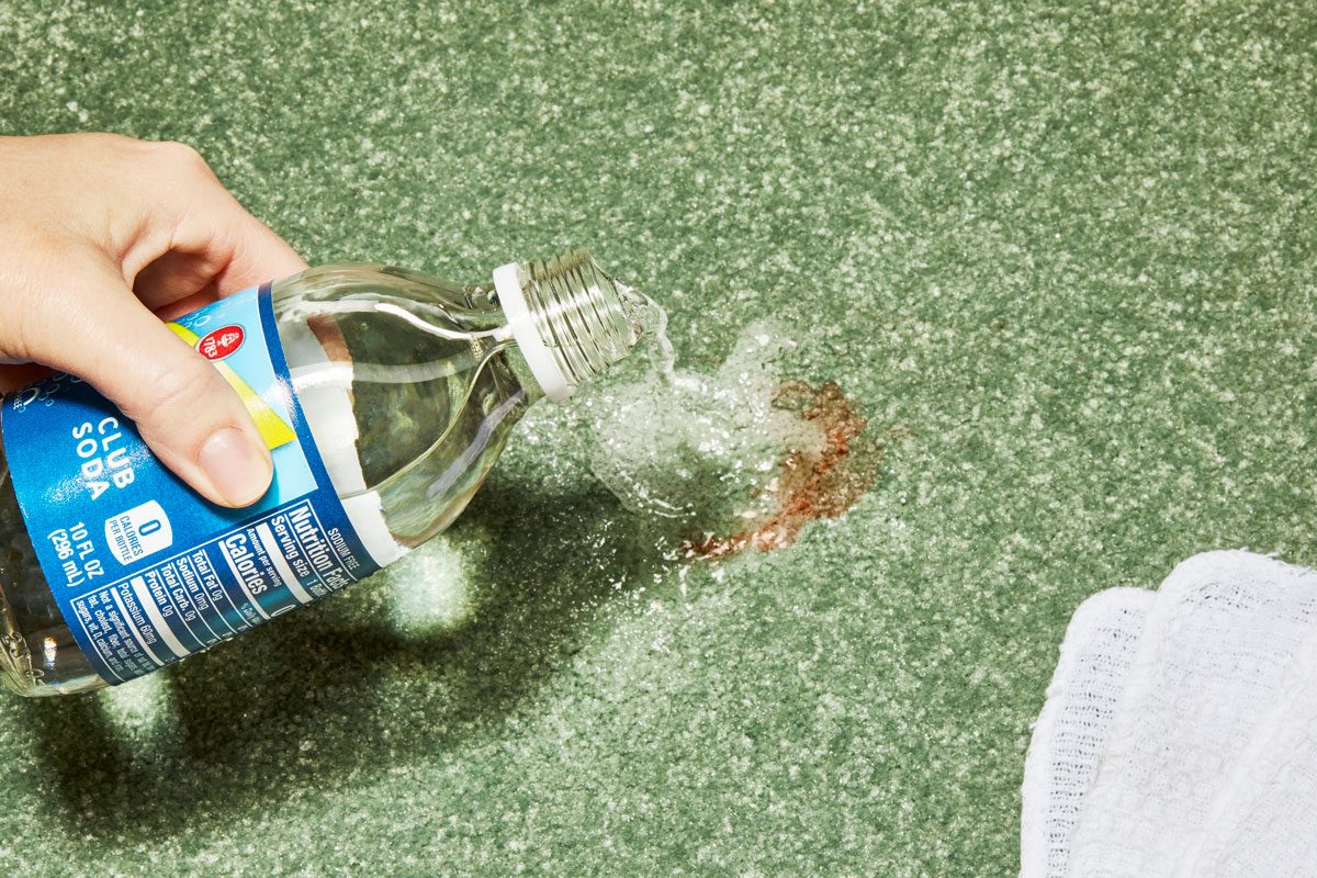 Hand pouring club soda on blood stain on green carpet with white cleaning cloth nearby