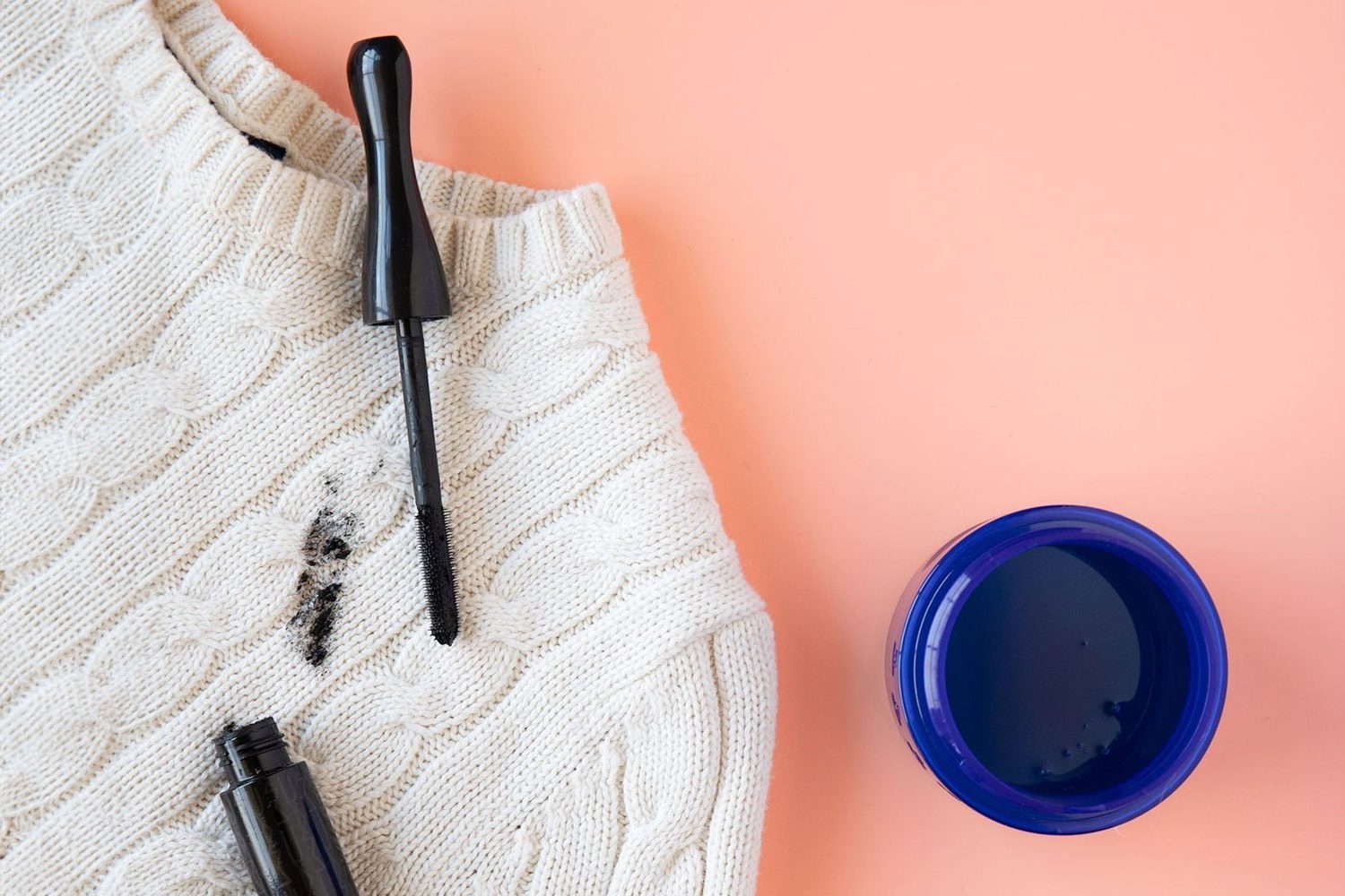 White cable knit sweater with a mascara stain next to a capful of laundry detergent on peach background