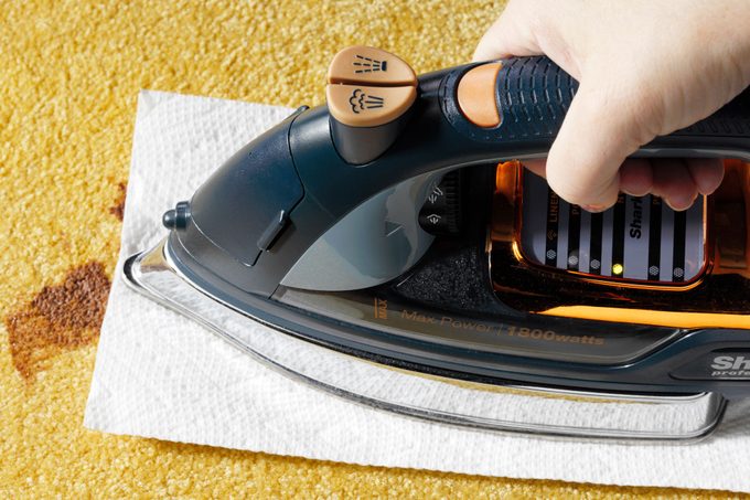 hand using an iron over a paper towel on the chocolate stain to remove it from the yellow carpet