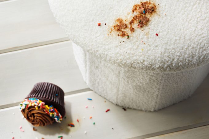 a chocolate stain on an upholstered stool with the fallen chocolate cupcake on the floor nearby