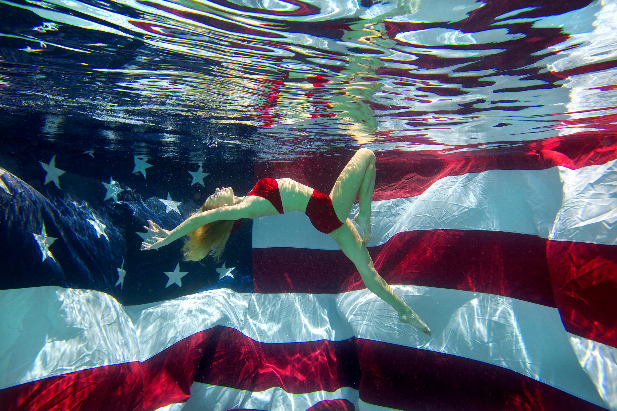 Portrait of a woman in a bikini underwater in a swimming pool with an American flag in the background in San Diego, California.