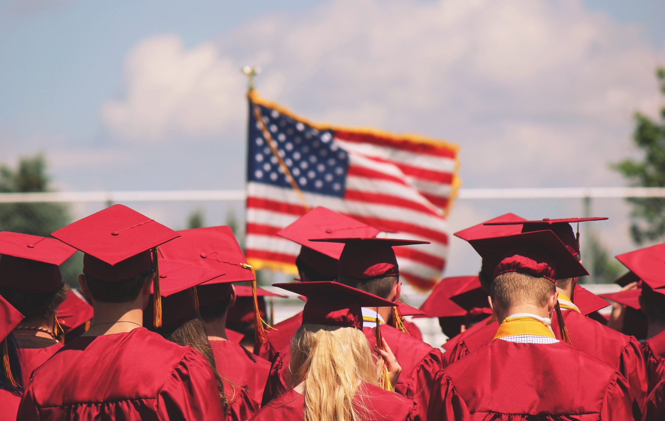 Rear View Of Graduates Standing Outside On A Sunny Day With An American Flag In The Background