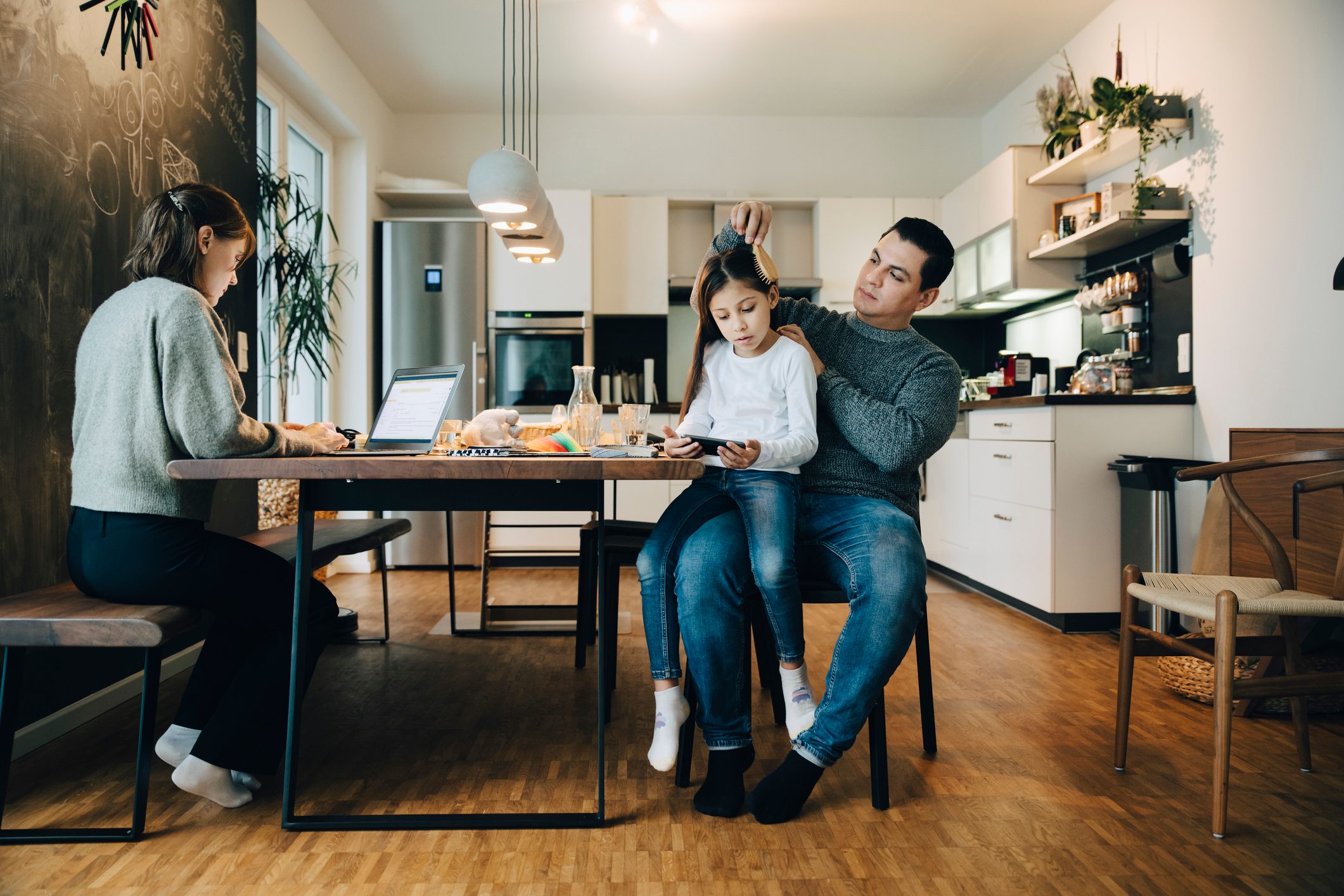 Father combing daughter's hair while mother working over laptop at home