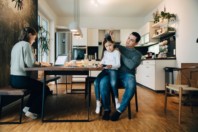 Father combing daughter's hair while mother working over laptop at home