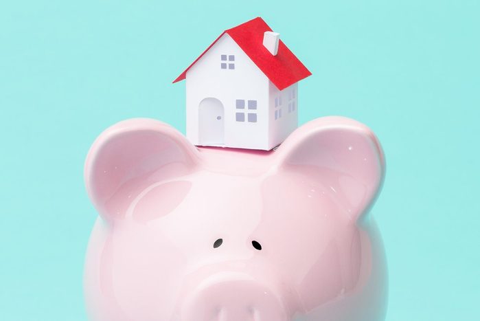 small house sitting atop a piggy bank against teal background