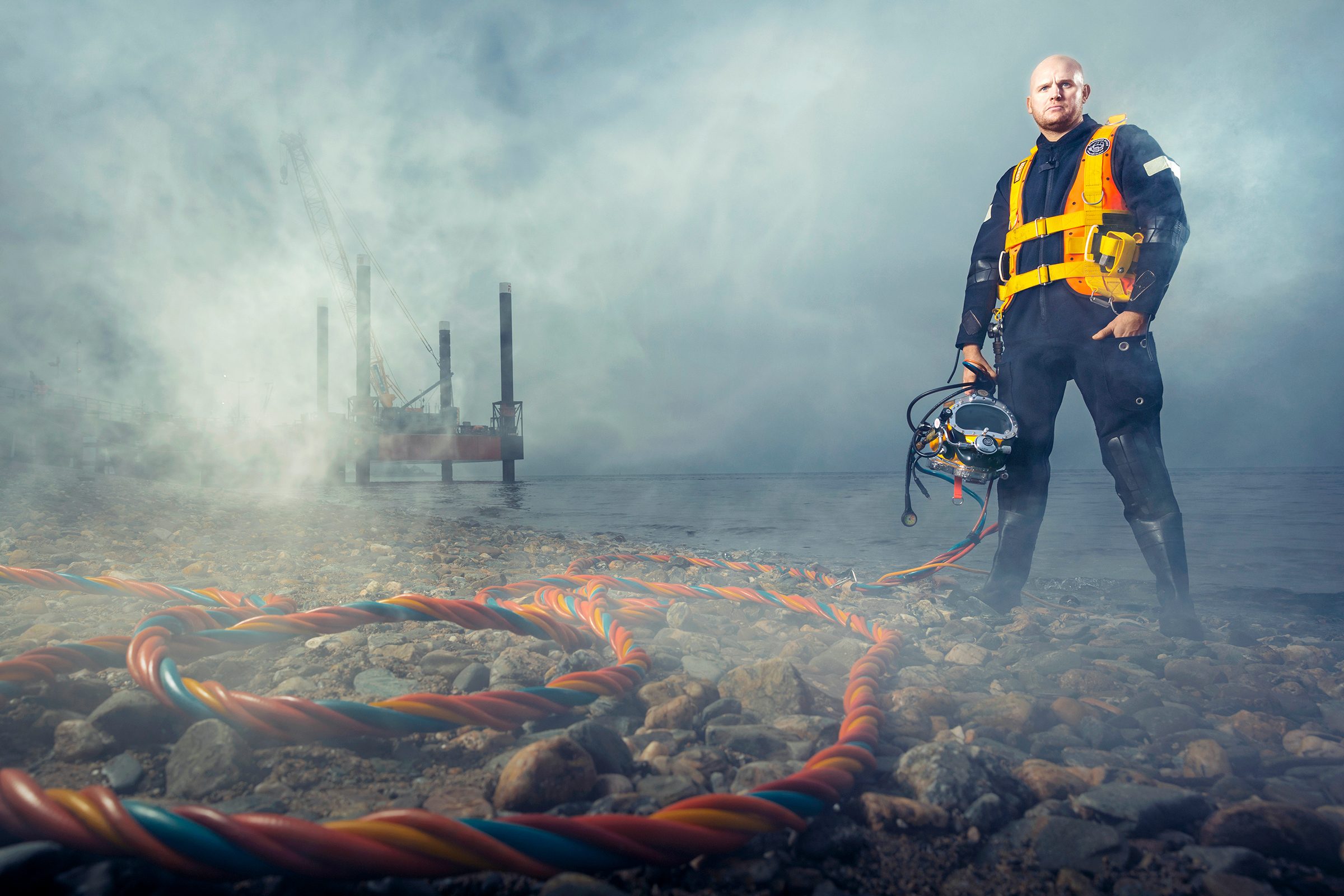 Chris Lemmons standing in deep sea diving gear in front of fog that shrouds background. Cords on the ground in the foreground