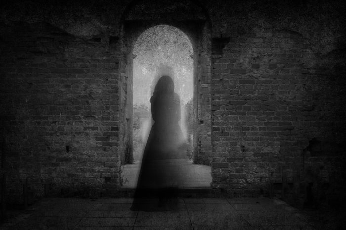 A spooky ghost of a woman in a dress, back to camera, framed by the archway of an old building. With a grunge, vintage, blurred edit.