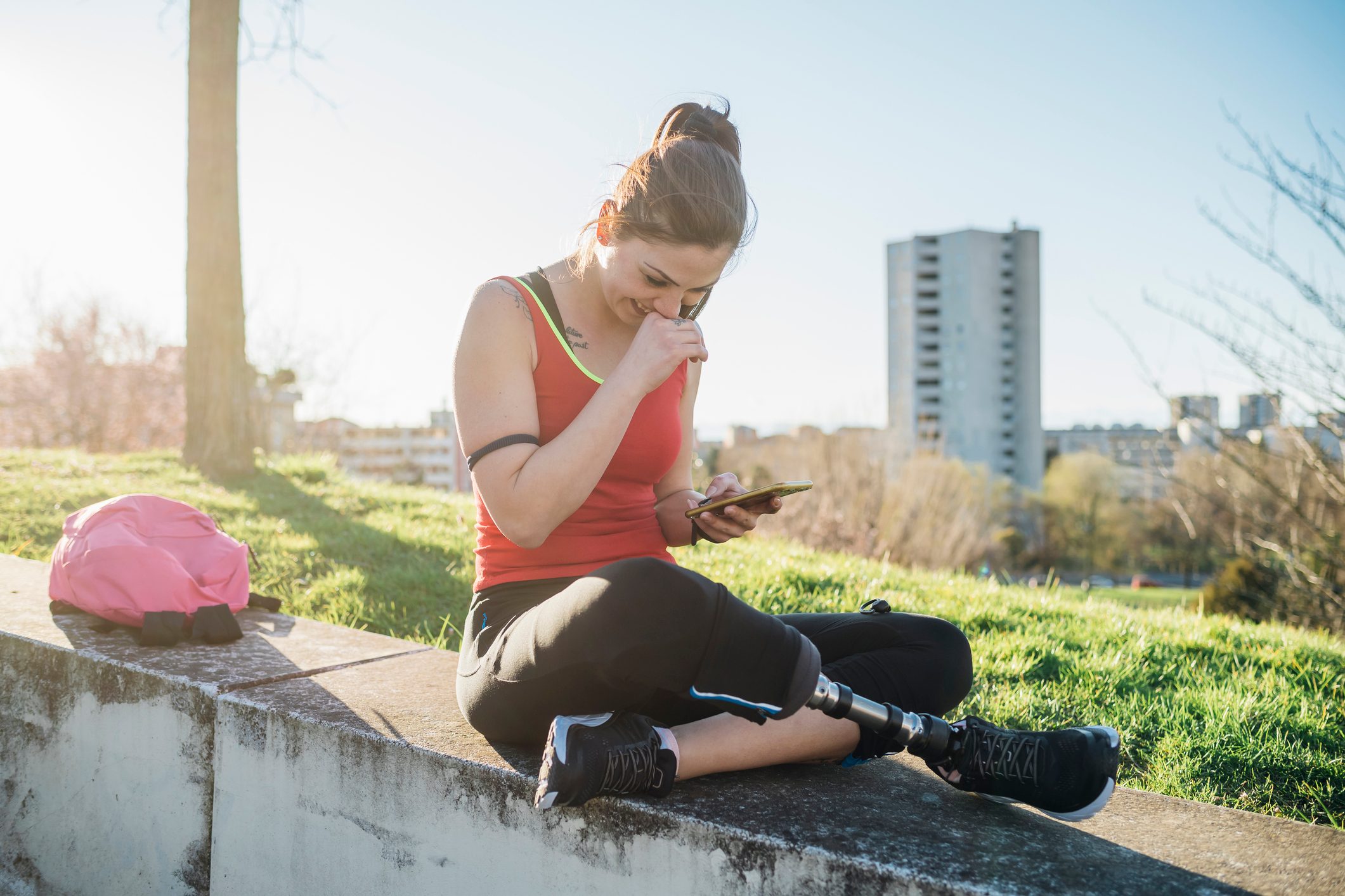 Sporty young woman with leg prosthesis sitting on a wall using smartphone to connect to social media