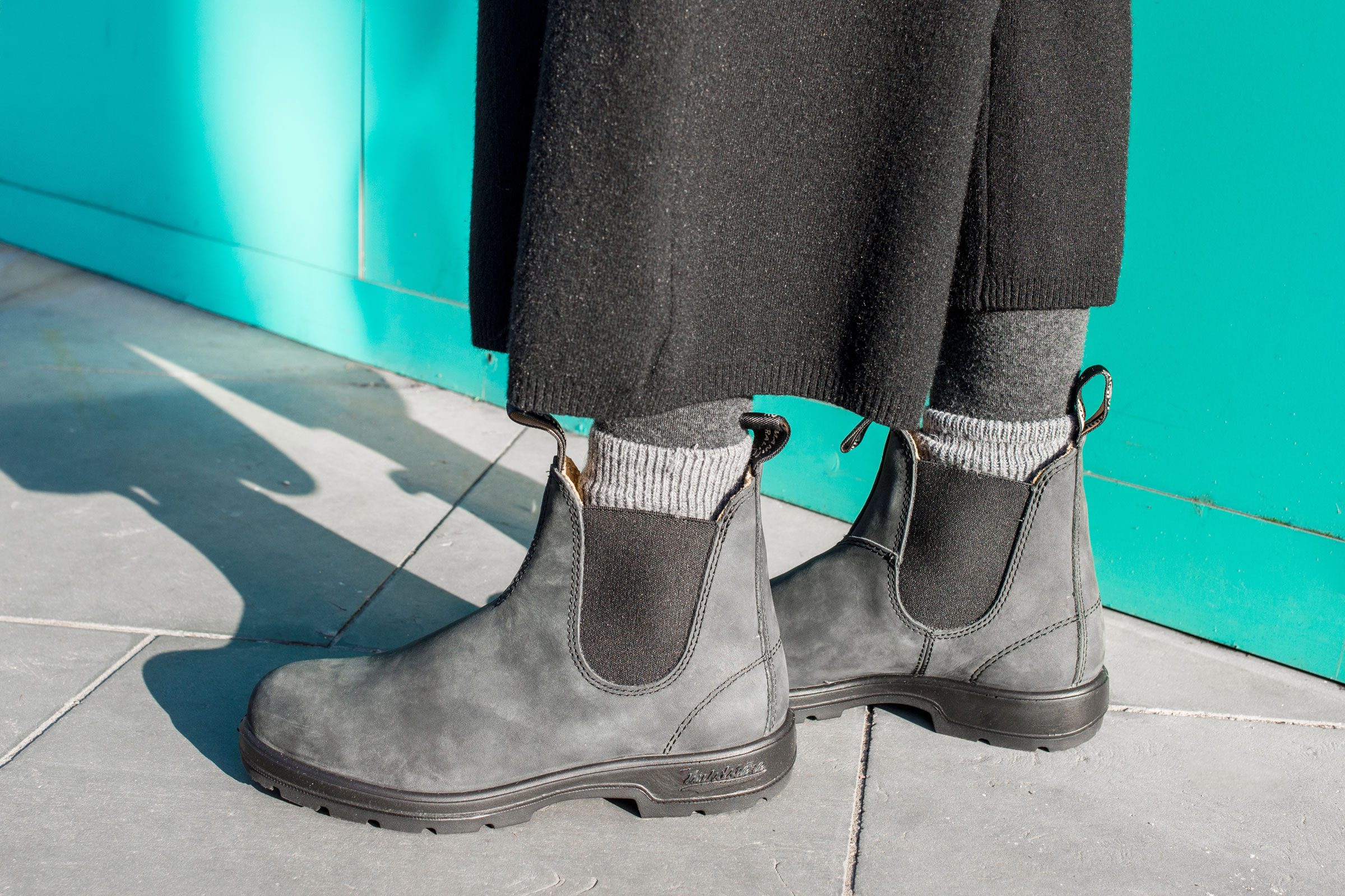 person wearing grey Blundstone Chelsea Boots outside on a sidewalk near a turquoise coloredwall