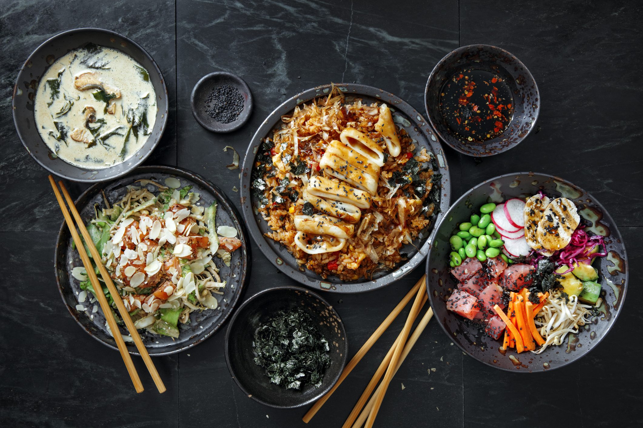 Tradition Japanese Dishes for Family Dinner on dark background. Japanese Grilled Squid with Teriyaki Sauce on Topped Fried Rice Bowl. Ahi Tuna Poke Bowl. Salmon and Avocado Salad with ginger dressing. Cream soup with eel.