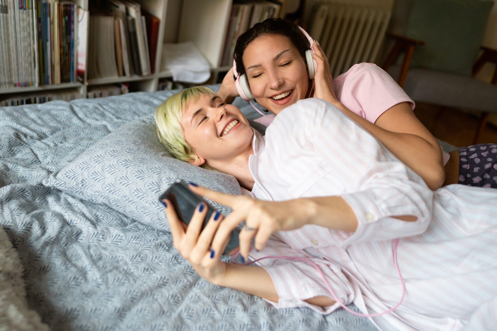 Smiling lesbian couple lying in bed together and listening to the music on a smartphone