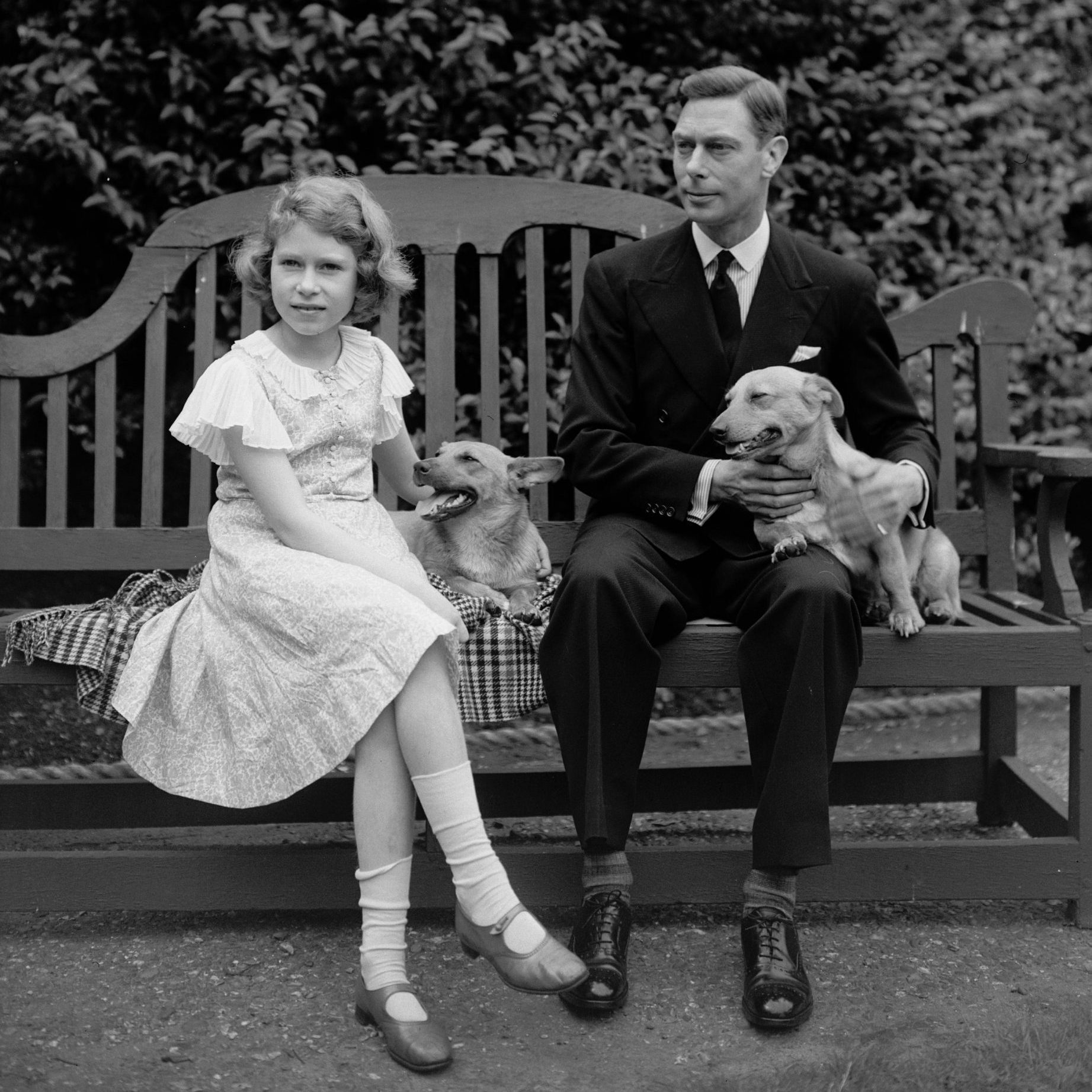 July 1936: George, Duke of York and Princess Elizabeth sitting on a bench with their corgi dogs in the grounds of their London home, 145 Piccadilly. (Photo by Lisa Sheridan/Studio Lisa/Getty Images)