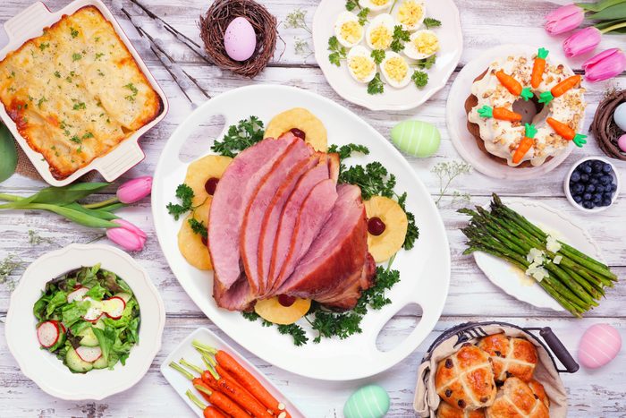 TraditionaTraditional Easter ham dinner. Top view table scene on a white wood background. Ham, scalloped potatoes, eggs, hot cross buns, carrot cake and vegetables.l Easter ham dinner. Top view table scene on a white wood background.