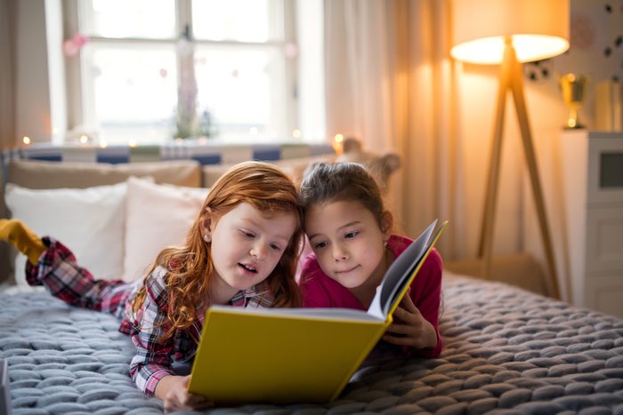 Two small girl friends lying on bed, reading book.