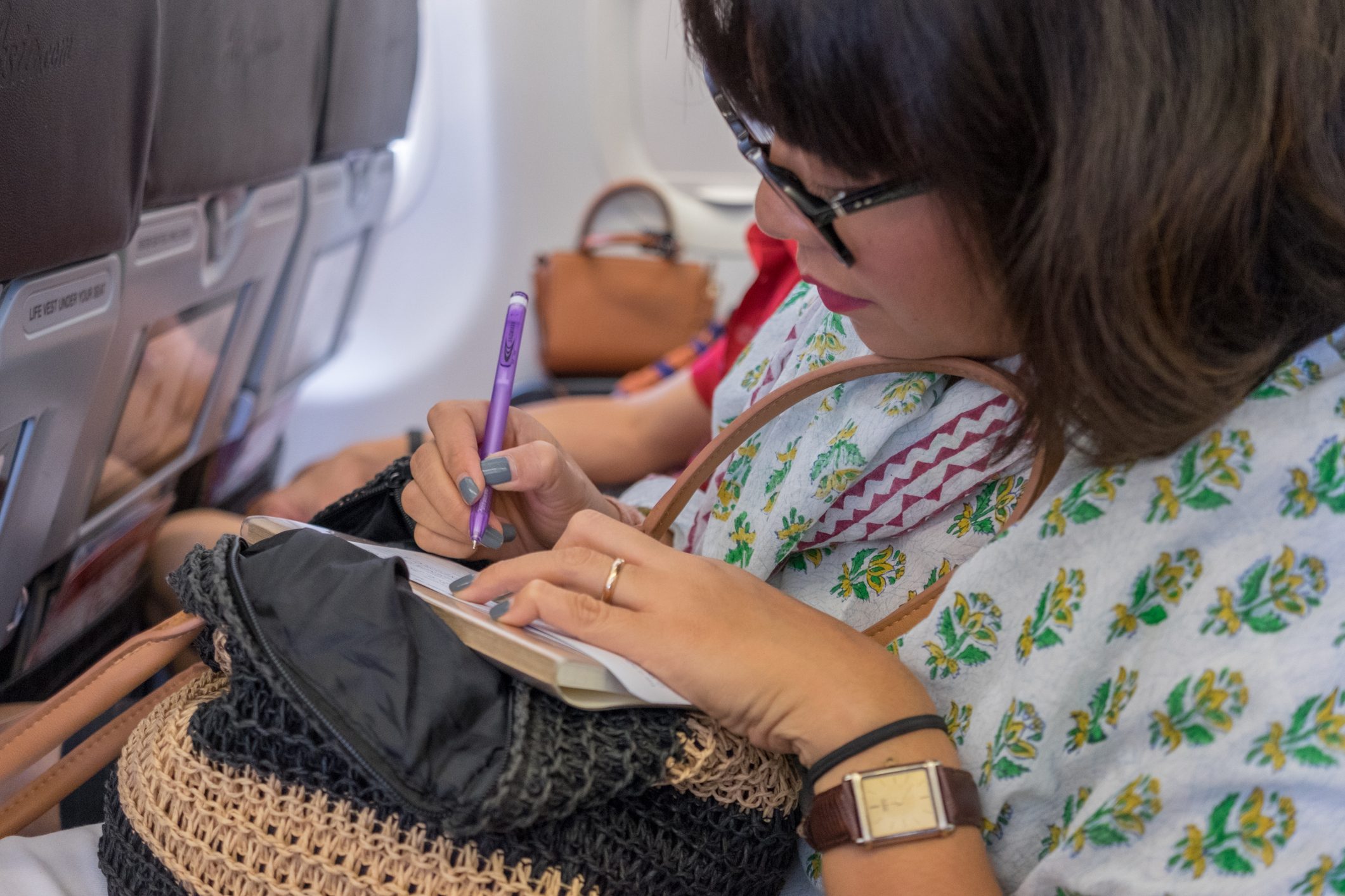 Asian woman with glasses seated on aircraft writing with pen.