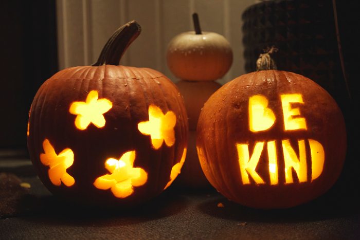Jack-O-Lantern pumpkins illuminated outside a house at night, one with flower cutouts and the other with the words Be Kind, Halloween decorations