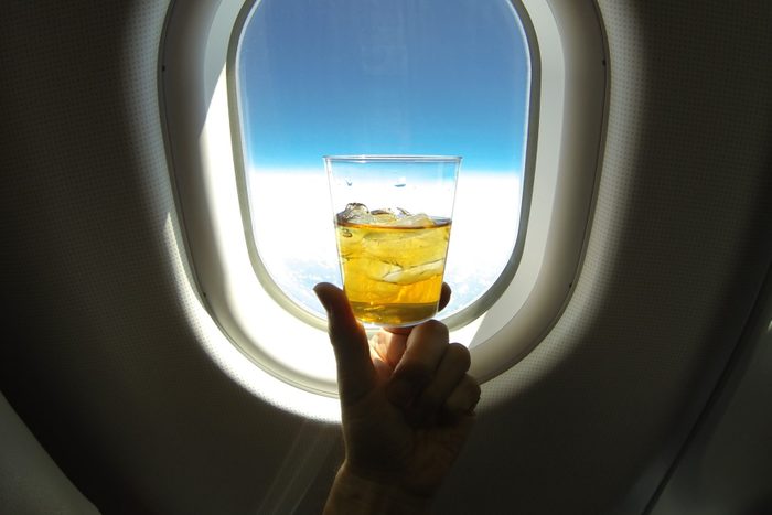 Refreshing glass of ice tea (or alcohol mix) centered on the window of a jumbo airplane, with blue sky above and snowy white clowds below.