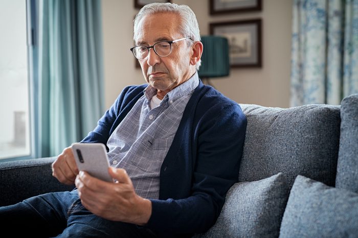 Serious senior man using smartphone sitting on the couch at home