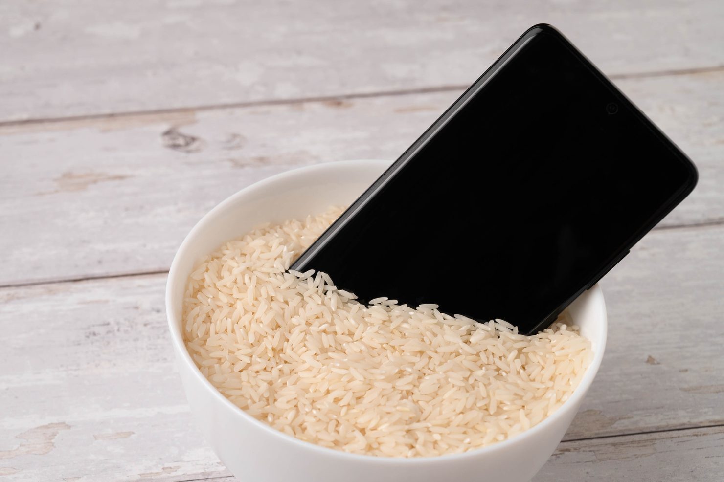 A mobile phone placed to dry in a bowl full of rice after the phone fell into water. Light wooden table background.