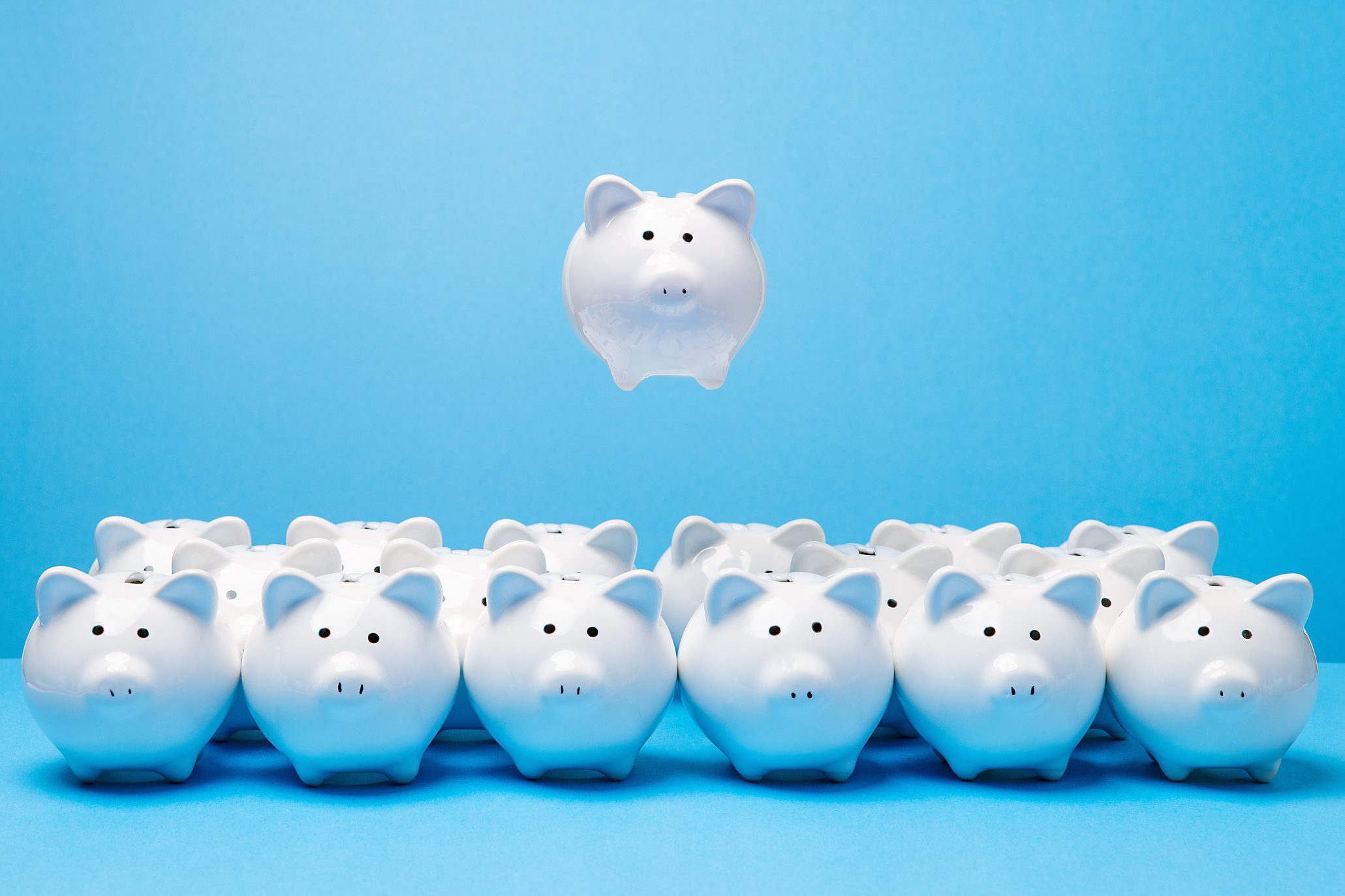 One little white ceramic piggy bank hovering in mid air above group of many other similar piggy banks facing the viewer on blue surface and background