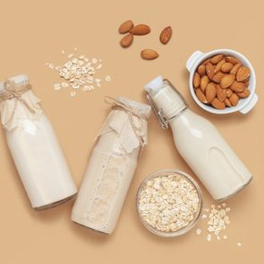 three glass jars of milk with almonds and oats to the side, on a tan background