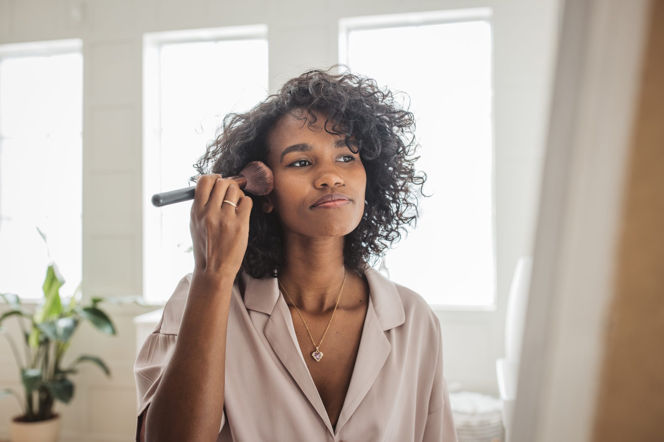 curly haired young woman applying foundation wit ha brush in the morning in her bathroom.