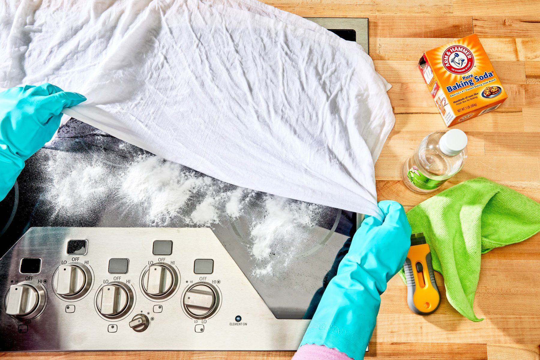 gloved hands covering glass top stove with hot towel, beneath the towel the baking soda on the stove top surface is visible.
