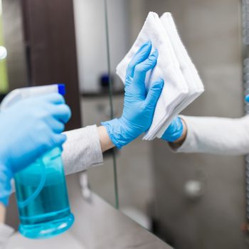 Close up of female hands with medical gloves polishing mirror with rag and using disinfectant spray. Housekeeping and cleaning service.