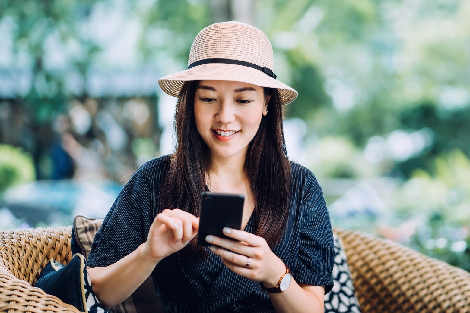 Beautiful young Asian female traveller with a straw hat surfing on the net while relaxing on lounge chair in an outdoor garden at luxury tropical resort