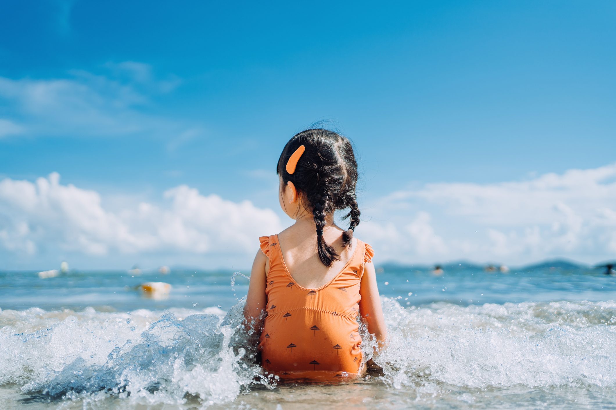 Rear view of lovely little Asian girl sitting by the seashore at the beach and being splashed by waves. Having fun at beach on a sunny Summer day